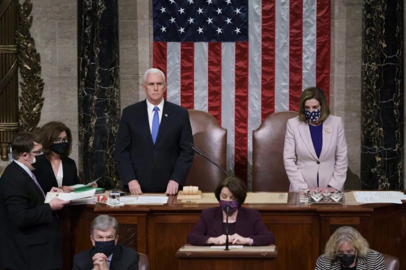 Vice President Mike Pence and Speaker of the House Nancy Pelosi stand on a podium in front of a large American flag. They are masked and solemn.
