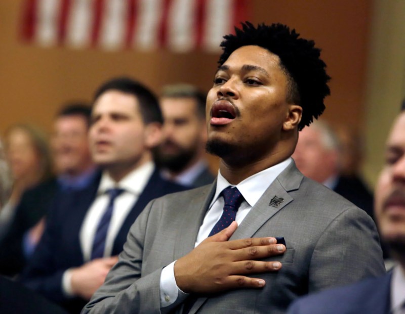 State representative Malcolm Kenyatta, a Black man, wears a gray suit and tie, and holds his right hand over his heart as he sings the National Anthem.