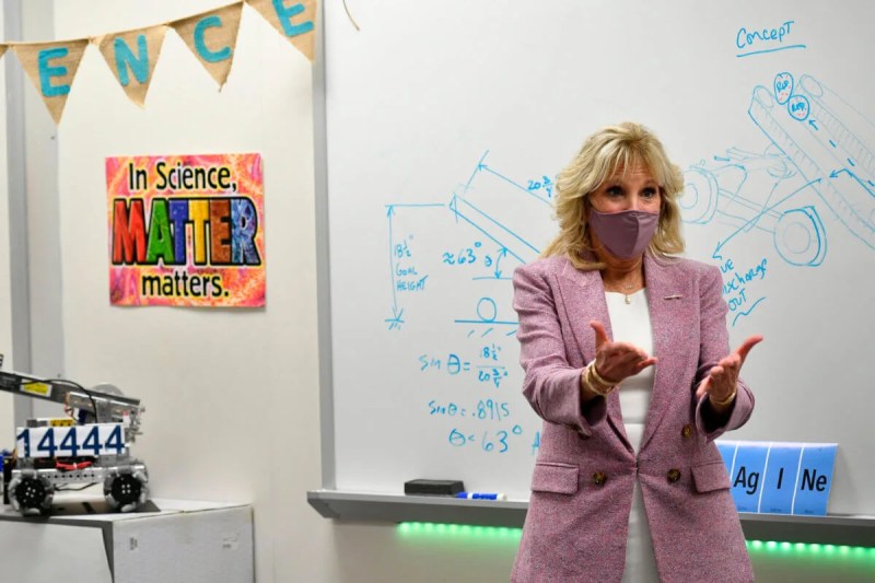 First lady Jill Biden stands in front of a white board in a robotics classroom. She is wearing a white dress, a dusky pink blazer, and a pink mask over her mouth and nose.
