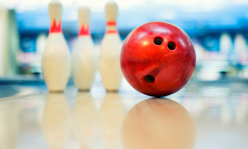 A red ball sits on a clean bowling lane with three bowling pins.
