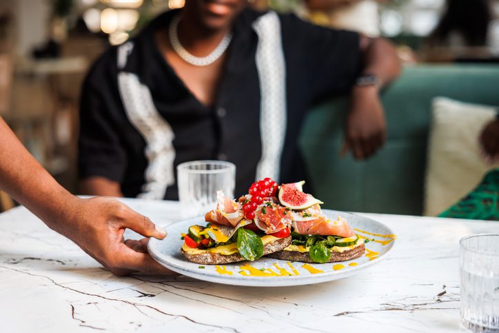 A hand is placing a plate of colorful bruschetta on a marble table in a chic Black-owned restaurant, with an out-of-focus figure in the background.