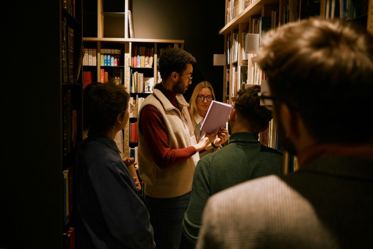Group of people standing in a dimly lit library, talking about a book.